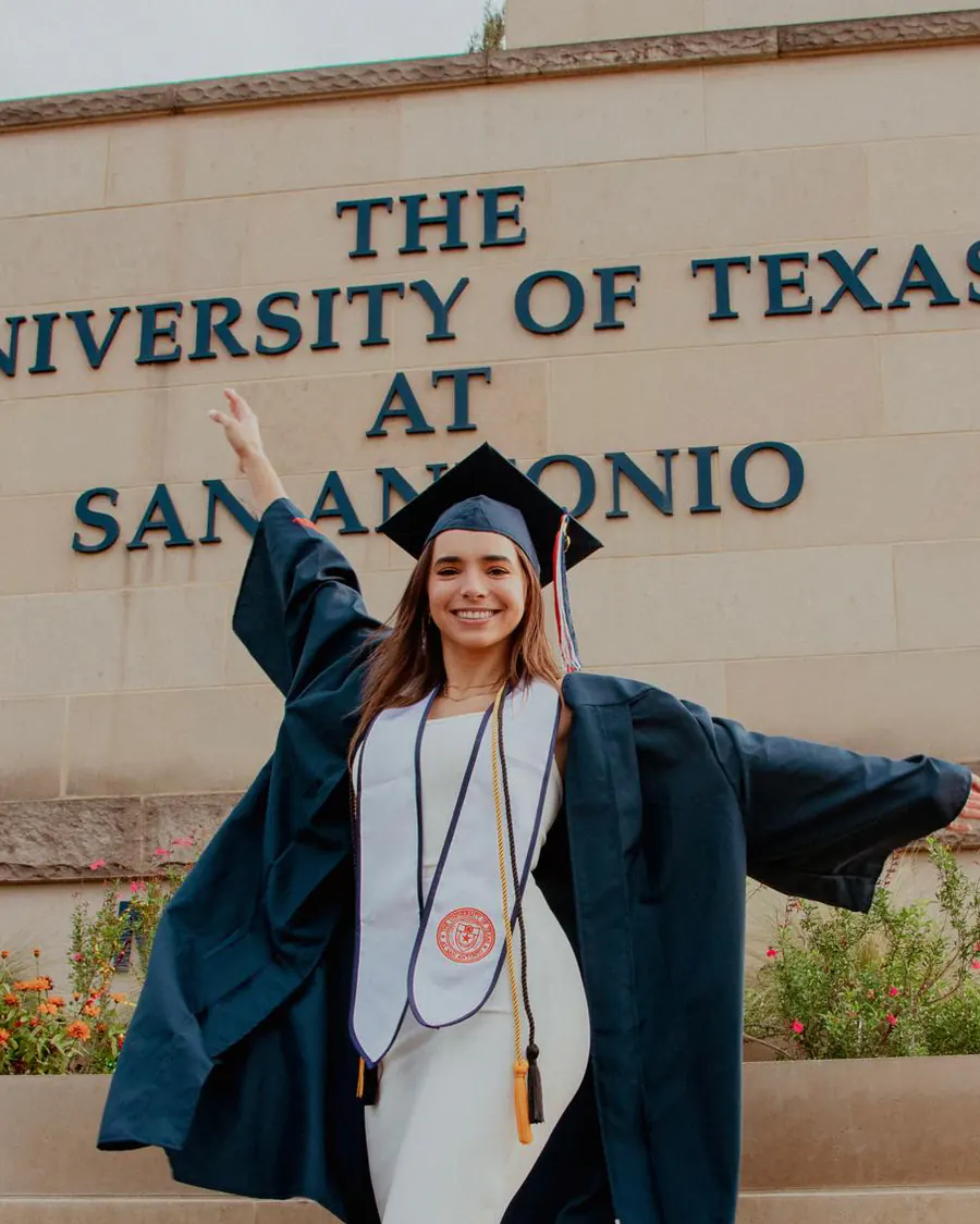 Natalia in cap and gown in front of the UTSA sign on graduation day