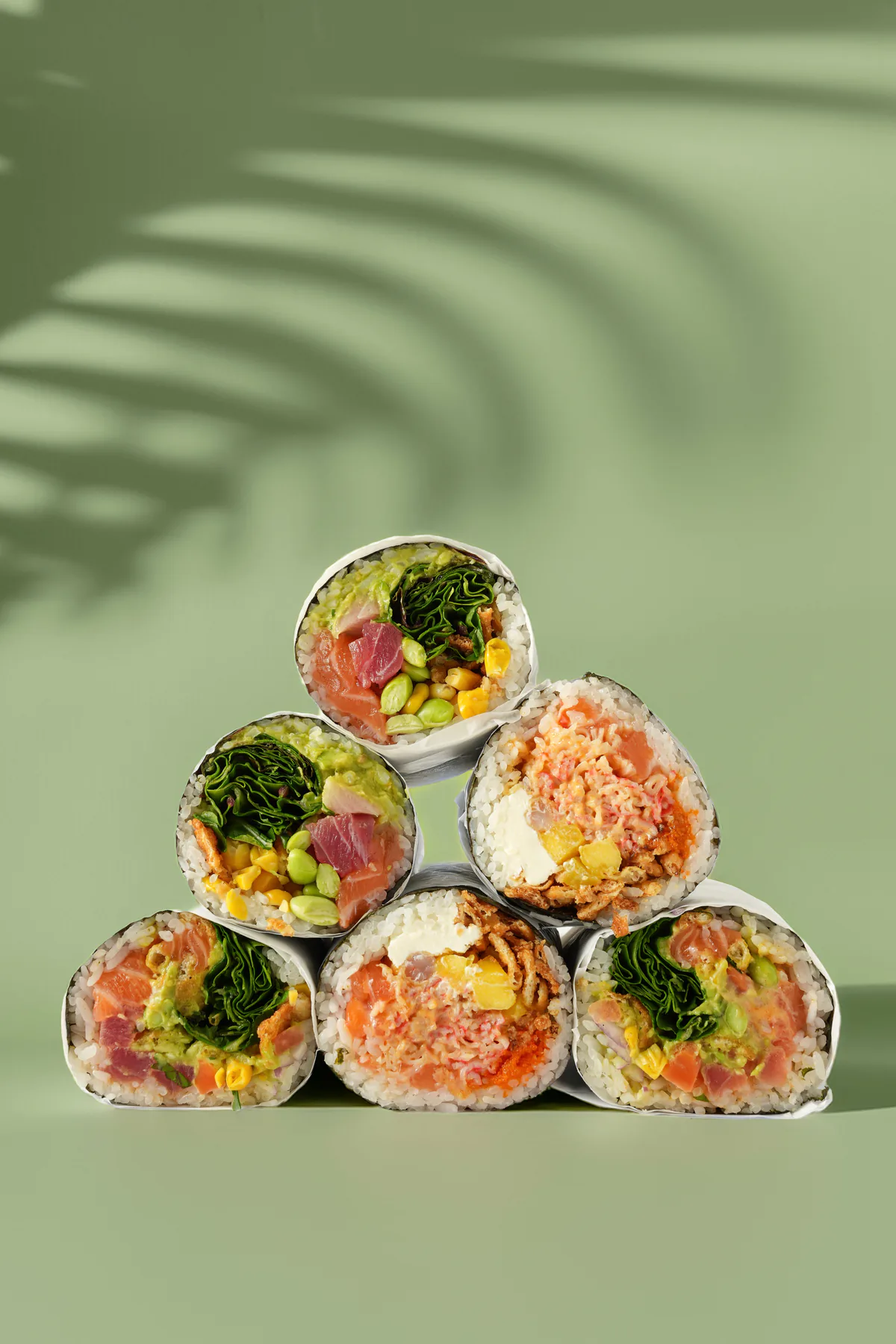 Six sushi burritos stacked in a studio shot with palm-leaf shadows